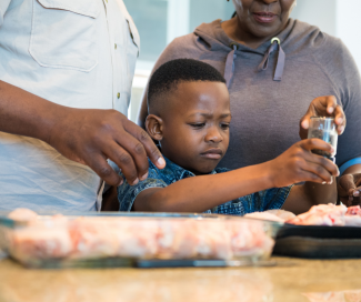 A little boy helps cook dinner with his grandparents by adding pepper to the dish. 