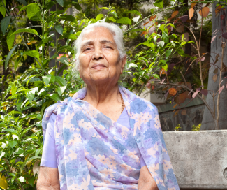 An older woman with a purple patterned shirt sits on a park bench with ivies behind her.