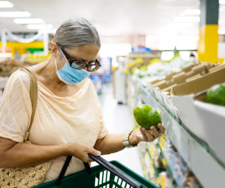 An older woman with a mask adds a green pepper to her grocery shopping basket.